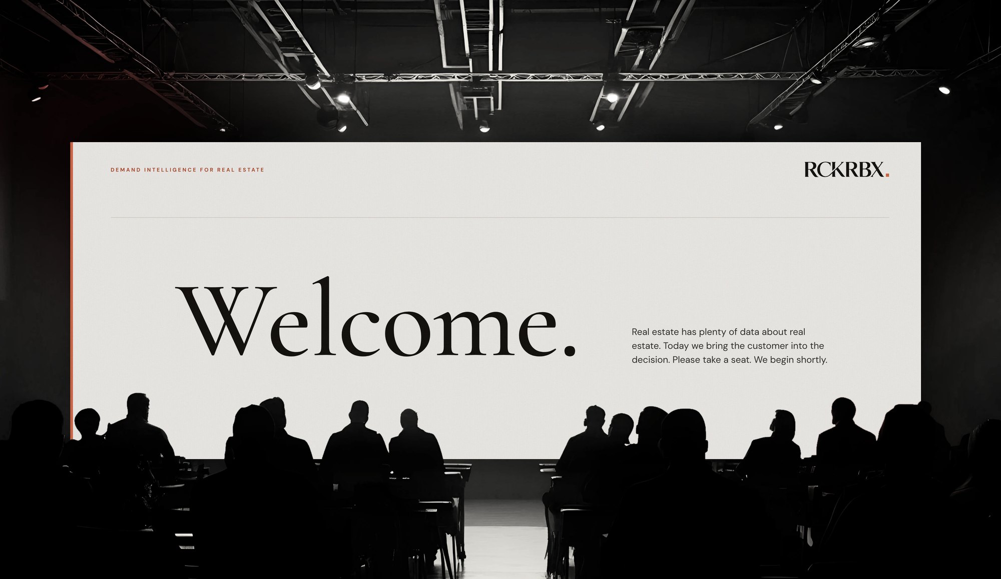 A conference screen reading Welcome in large serif type, an audience in silhouette, the RCKRBX wordmark top right