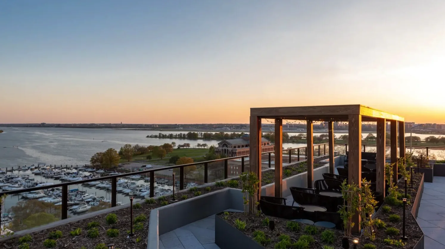 Photograph from the rooftop at sunset: a pergola and lounge seating above the marina, with the Anacostia and Fort McNair beyond