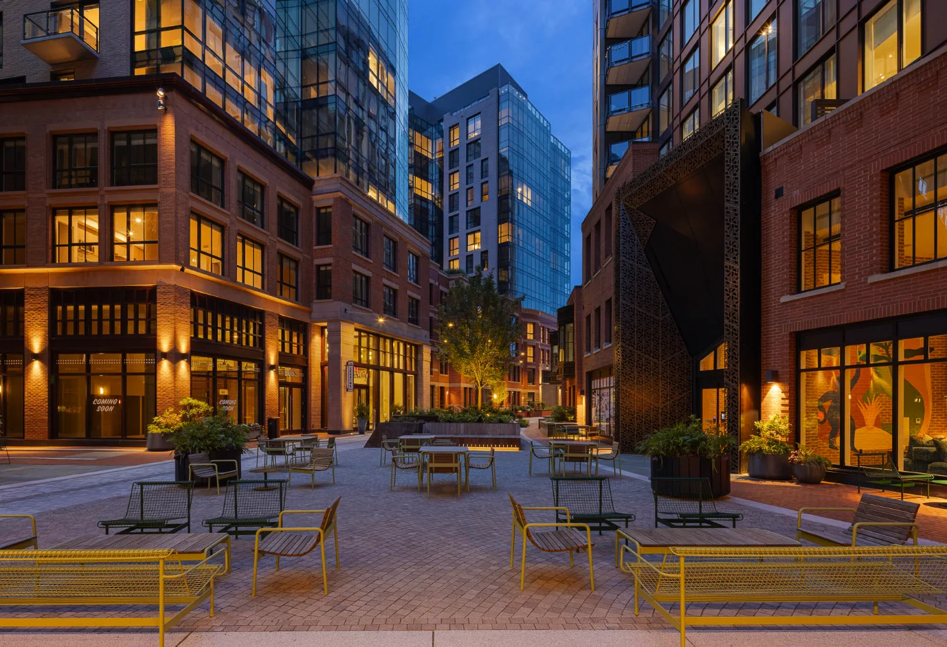 Photograph of the plaza at Everly at The Stacks at dusk: yellow benches and young trees between a brick building and a glass tower, storefronts not yet open