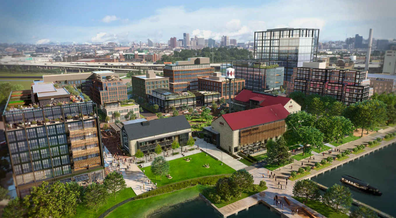 Aerial rendering of Port Covington: two restored warehouses with red roofs on the waterfront green, new apartment and office blocks behind, the Baltimore skyline beyond