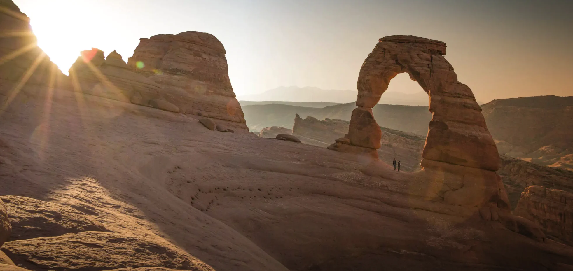 Delicate Arch at sunrise with two figures beneath it and the La Sal mountains beyond