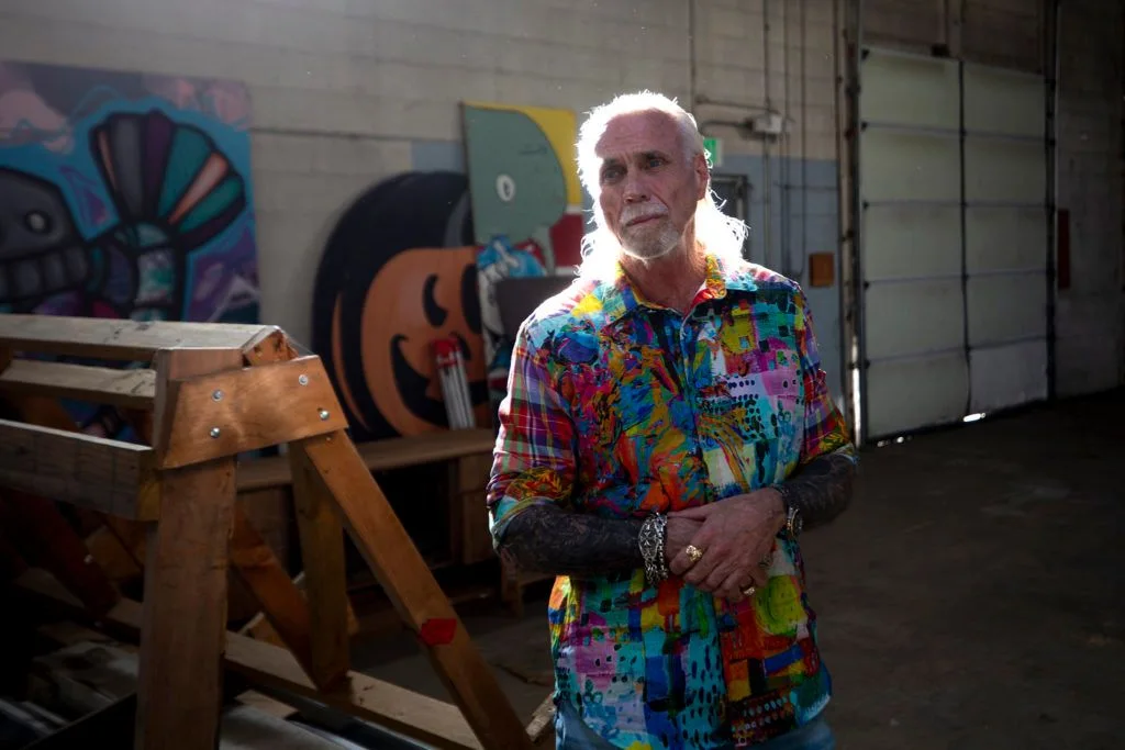 Photograph of Bernard Hurley in a patterned shirt, standing in a RiNo warehouse with large painted panels behind him