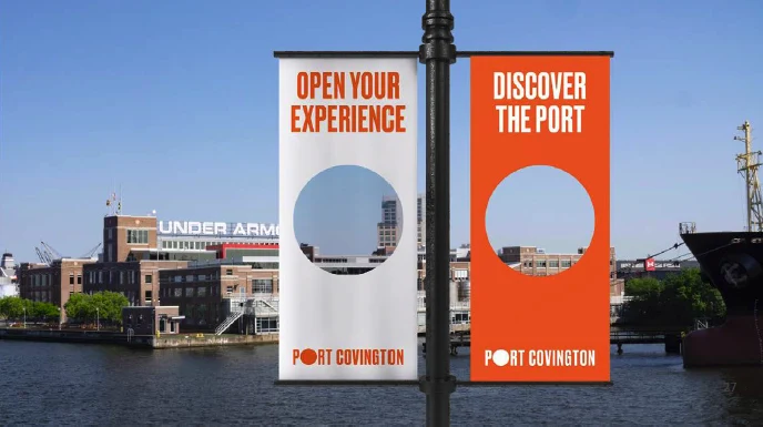 Two orange street banners on a lamp post by the harbor: Open Your Experience and Discover the Port, each with a circular window