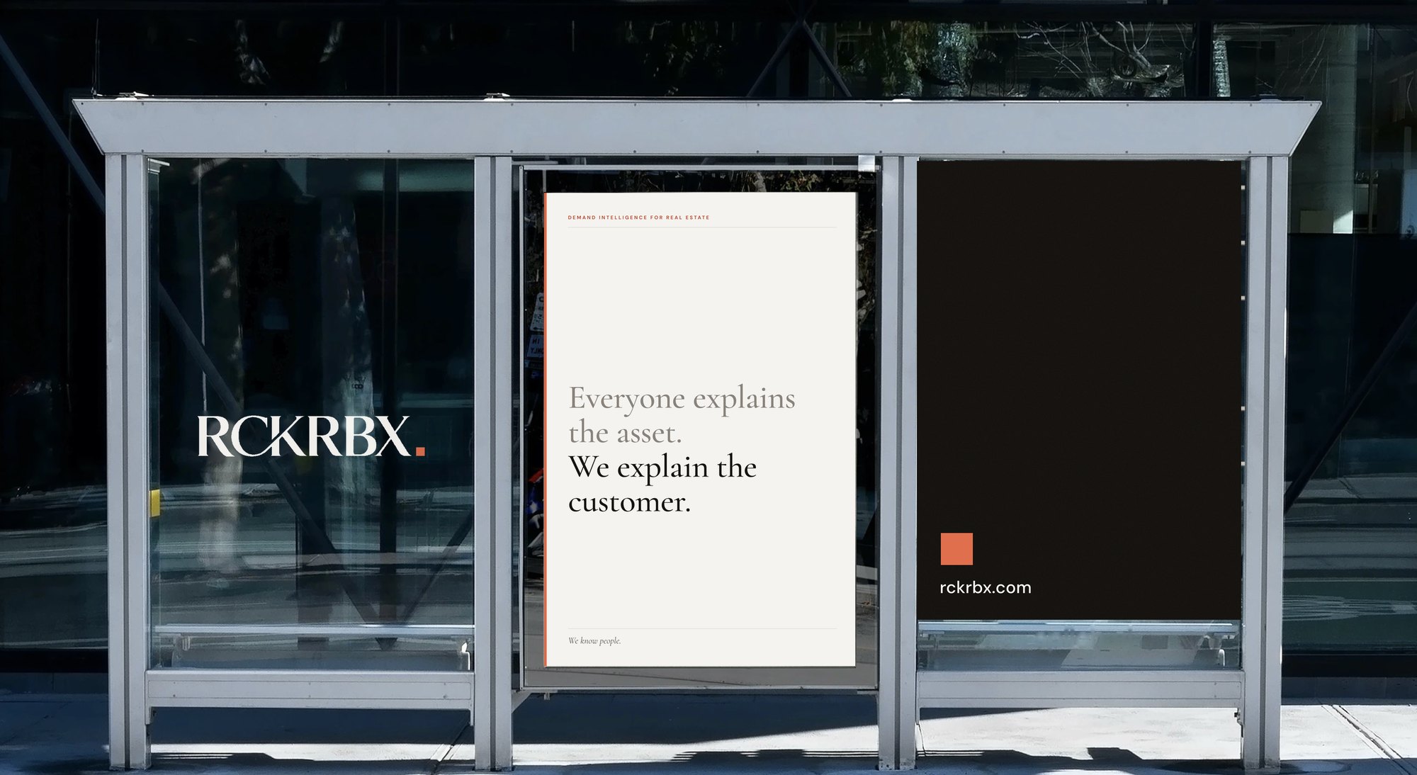 A bus shelter with three RCKRBX panels: the wordmark on glass, a cream poster reading Everyone explains the asset. We explain the customer, and a dark panel with the orange square and rckrbx.com