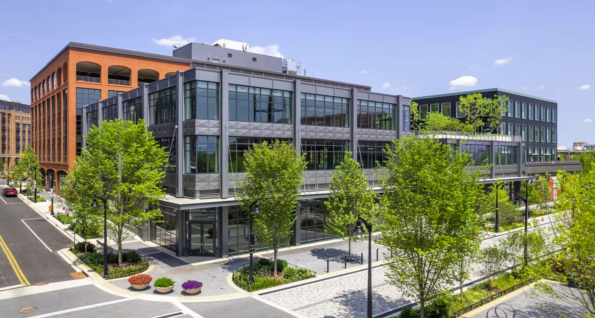 Photograph of the built Rye Street Market block: a glass and steel office building with a brick parking structure behind, young trees and planters on a paved plaza