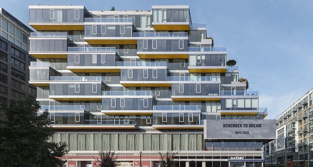 Photograph of the built building: stacked glass volumes stepping back from the street, yellow soffits, a Remember to Dream billboard on the retail base