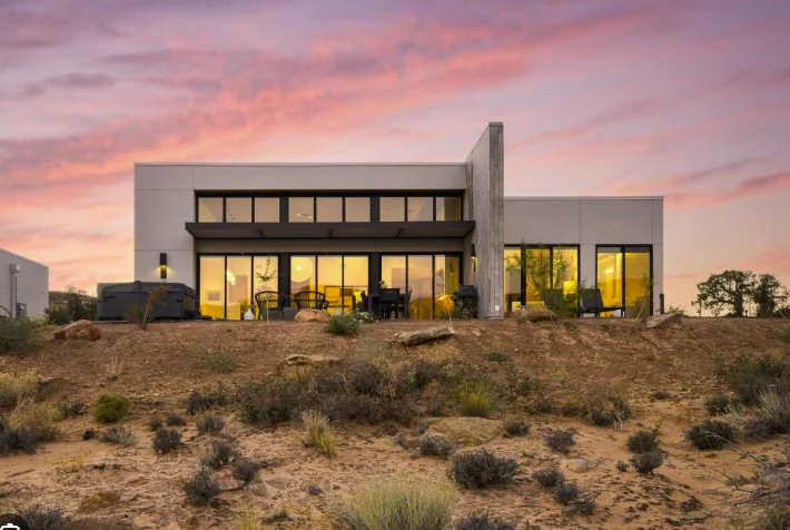 Photograph of a built casita at dusk: a flat-roofed stucco house with a lit glass wall and a hot tub, on a rise of red earth and sage